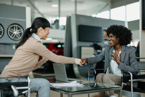 Two women shaking hands across a dealership desk closing a car purchase representing successful MSRP negotiation and taking advantage of January car deals and end of year car deals